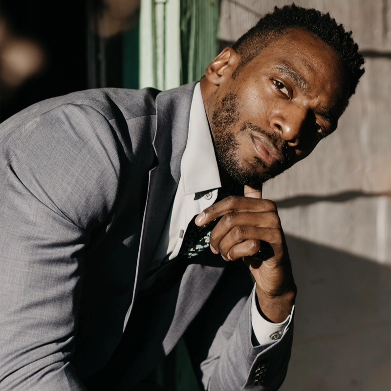 Daniel J. Watts, in a light gray suit and white shirt, poses thoughtfully with his chin on his hand. His short hair and trimmed beard catch the sunlight, casting shadows across his face against a blurred green and beige background.
