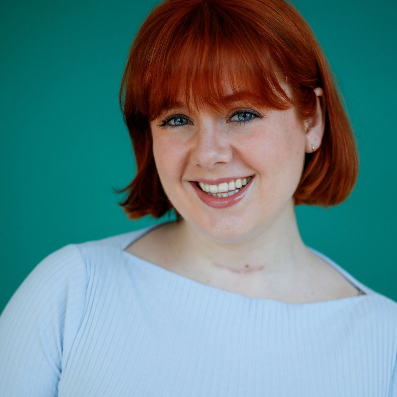 Emily J. Whalen, a young woman with short, straight red hair and bangs, smiles warmly. She wears a light blue ribbed top and small earrings against a solid teal background, creating a bright and cheerful atmosphere.