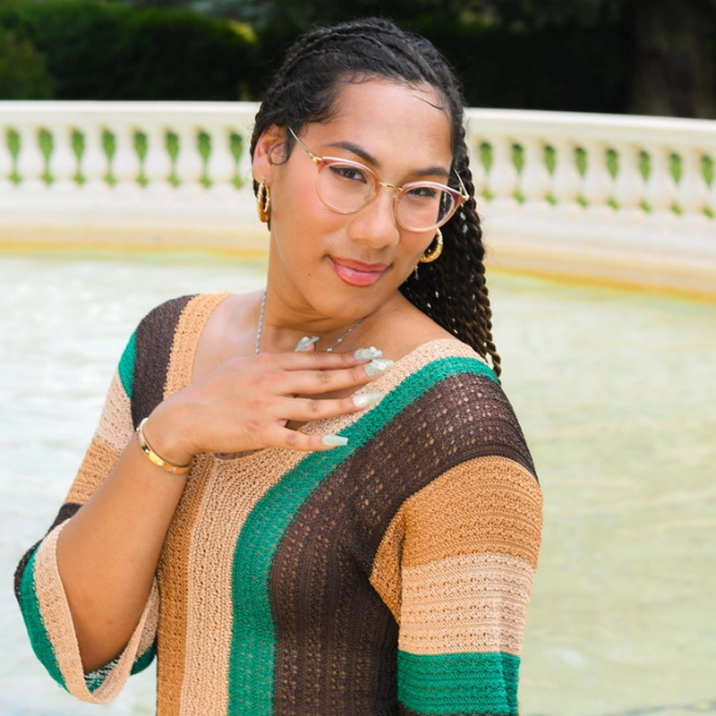 Shawn Williams, with braided hair, clear glasses, and hoop earrings, stands outside by a light-colored fountain. They wear a striped knit top in tan, brown, and teal, long nails, and a gold bracelet while gently touching their chest.