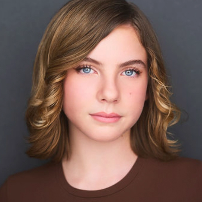 Jake Hamilton, a young person with shoulder-length, wavy light brown hair and blue eyes, gazes calmly at the camera. They have fair skin and wear a brown top. The plain, dark background keeps the focus on their face.