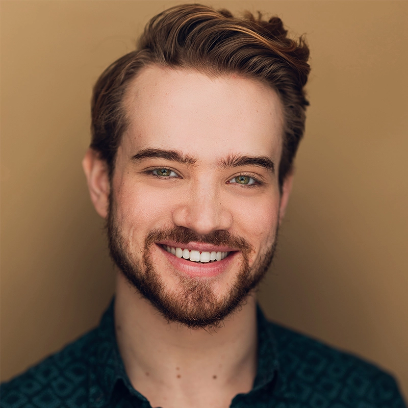 A young man with light skin, short brown hair swept to the side, and a trimmed beard smiles warmly. He has green eyes and is wearing a dark patterned shirt. The background is a solid, warm beige color.