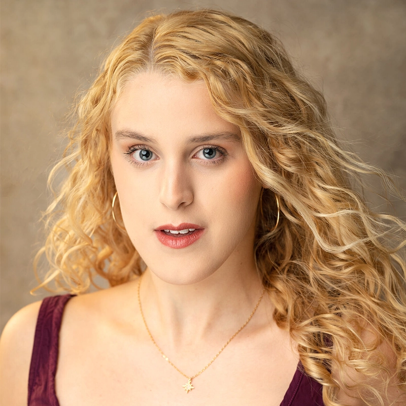 A woman with long, curly blonde hair poses against a neutral brown background. She wears a sleeveless burgundy top, gold hoop earrings, and a delicate gold necklace with a star-shaped pendant. Her expression is calm and confident.