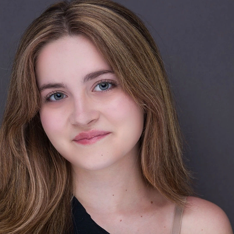 A young woman with fair skin, long light brown hair, and blue eyes smiles softly at the camera. She wears a sleeveless top with one black strap and one beige strap, against a plain gray background.