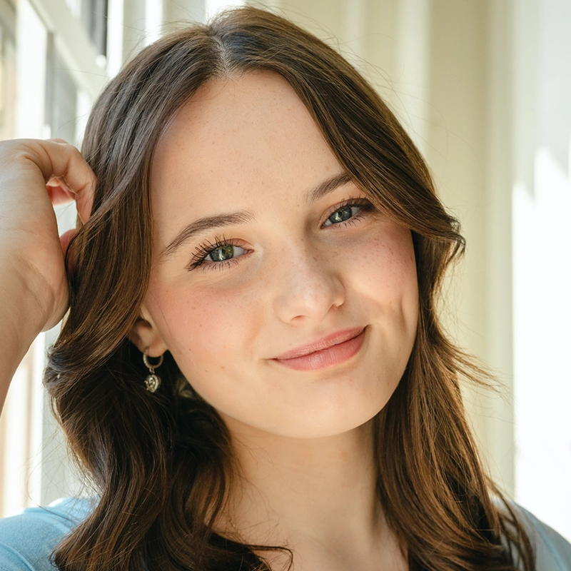 A young woman with long brown hair and green eyes smiles gently indoors. She has light skin, subtle makeup, and is wearing earrings. She touches her hair with one hand. Sunlight streams through large windows behind her, creating a soft, bright background.