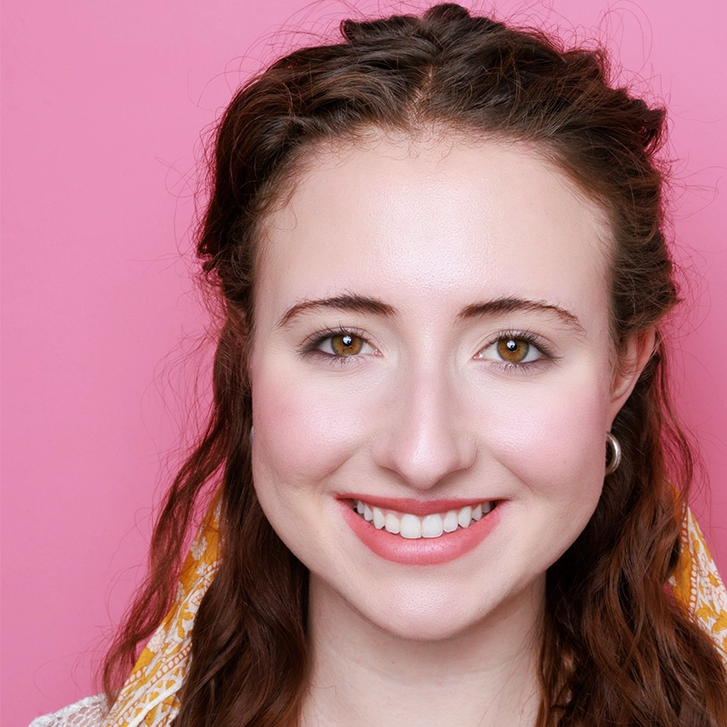 A young woman with fair skin, wavy brown hair, and hazel eyes smiles at the camera. She wears a yellow-patterned scarf and silver hoop earrings. The background is solid pink. Her makeup is natural and her hair is parted in the middle.