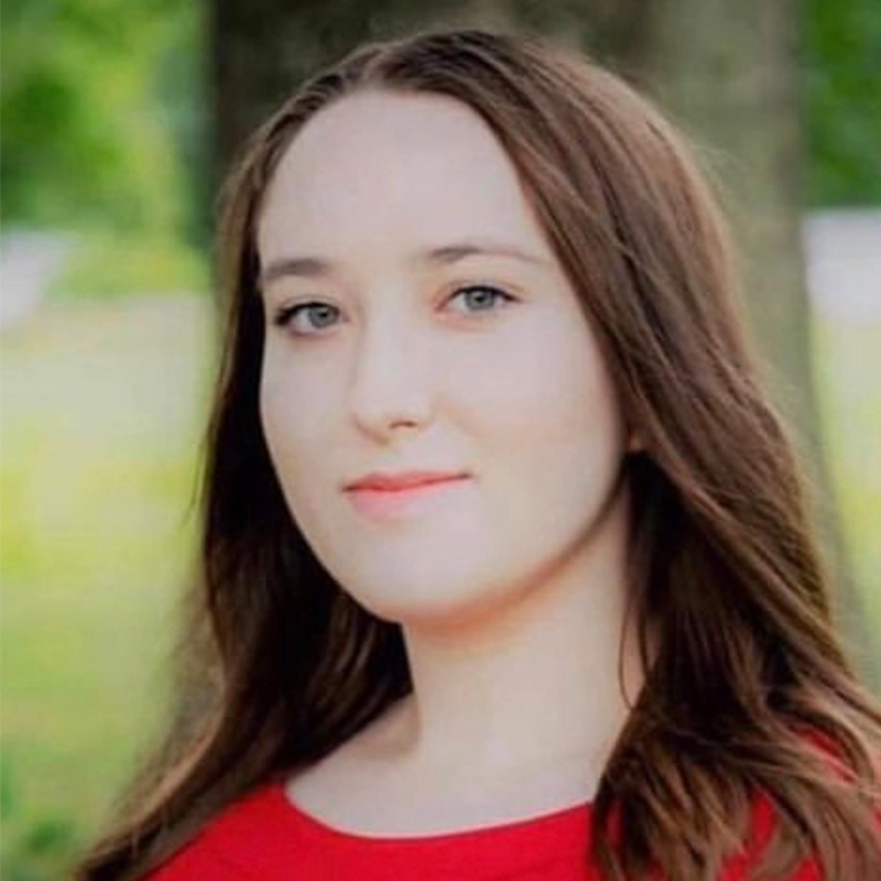 A young woman, Margaret Kelly, with light skin and long brown hair poses outdoors in front of blurred green trees. She has blue eyes, a slight smile, and wears a red top. The natural lighting creates a soft, warm atmosphere.