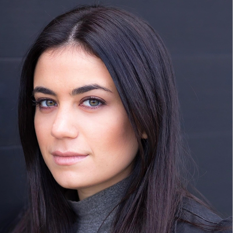 A woman with straight, long dark brown hair and light skin looks at the camera with a neutral expression. Channeling a Peter Salett vibe, she has green eyes, subtle makeup, and wears a gray turtleneck against a plain, dark background.