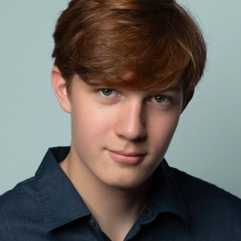 A teenage boy with fair skin and straight, light brown hair looks at the camera and smiles slightly. He is wearing a dark blue collared shirt. The plain, light greyish-blue background and soft lighting evoke the portrait style of Arielle Ziff.
