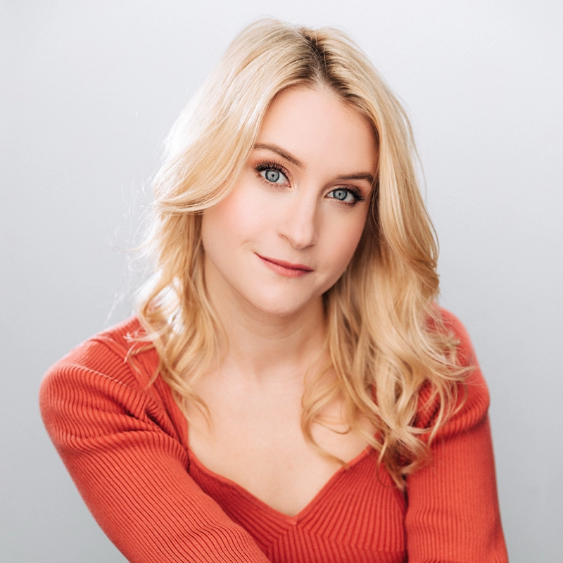 A woman with long, wavy blonde hair and blue eyes looks at the camera, smiling softly. She wears a ribbed, red-orange top with a v-neckline. The background is plain and pale, with soft lighting highlighting her face.