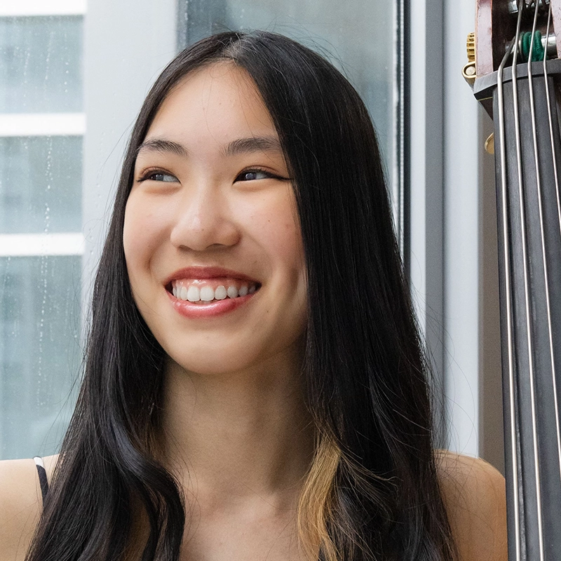 A young woman with long, straight black hair and a streak of blonde smiles brightly, revealing dimples. She stands indoors by a window on a rainy day. Part of a double bass instrument is visible beside her.