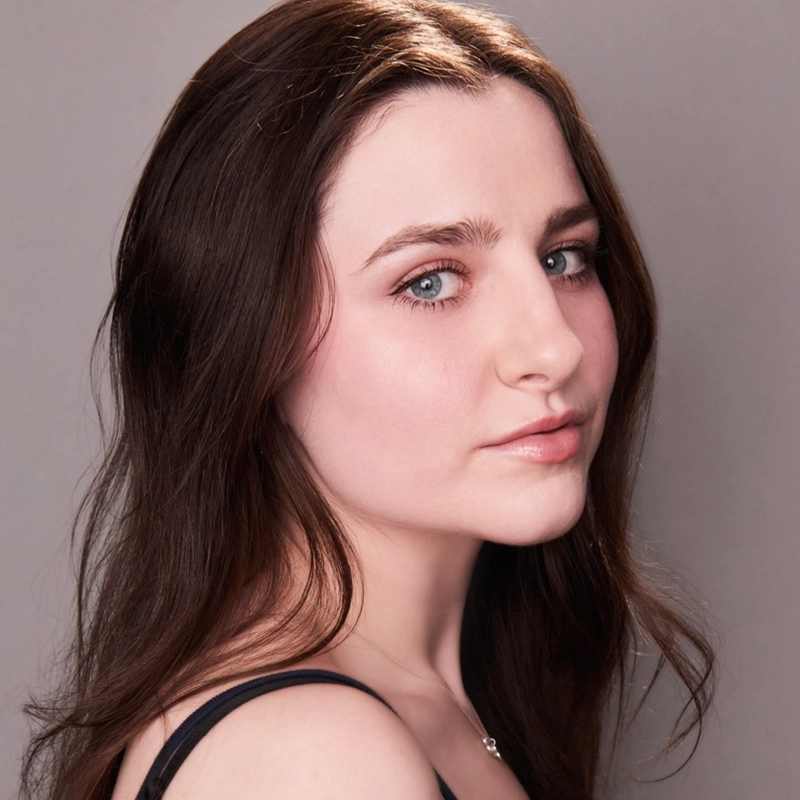 Arielle Ziff, a young woman with long brown hair and blue eyes, gazes softly at the camera. She has light skin, subtle natural makeup, and wears a black top with a silver necklace against a plain light gray background.
