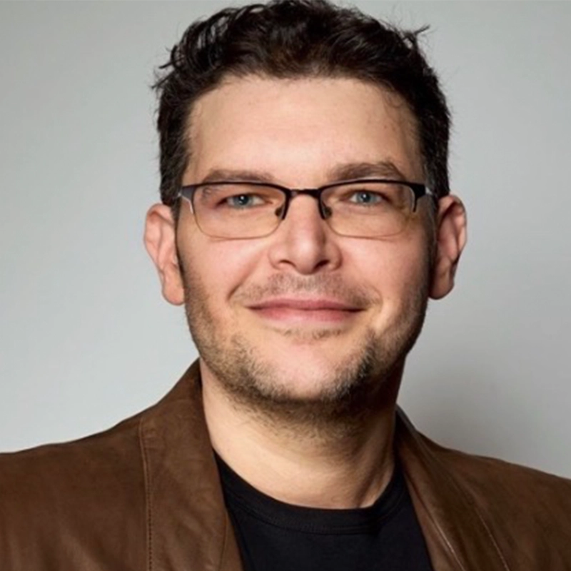 Peter Salett, a man with short dark hair and glasses, smiles at the camera. He is wearing a brown leather jacket over a black shirt. The background is plain and light gray.