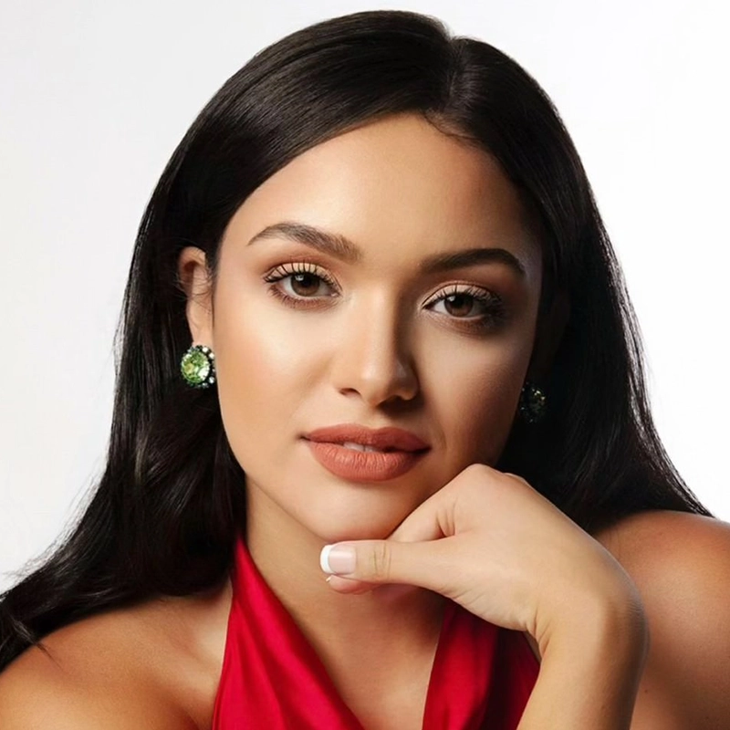 A woman with long, dark hair and smooth skin poses against a plain white background. She wears a red halter top, green gemstone earrings, and natural makeup. Her hand rests under her chin, and she gazes directly at the camera with a calm expression.