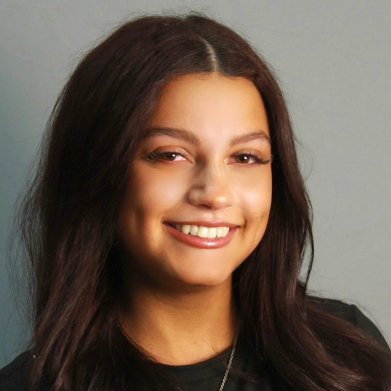 A young woman with long dark hair and a center part smiles at the camera. Annalisa Zambrano has dimples, wears natural makeup with winged eyeliner, and is dressed in a black top against a plain light gray background.