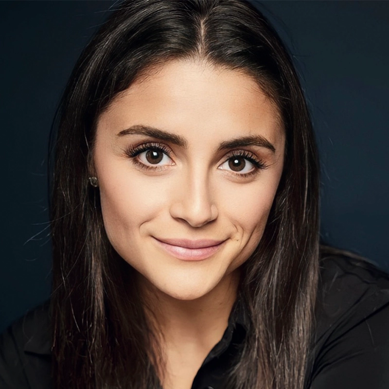 A young woman with straight, dark brown hair and brown eyes smiles softly at the camera. She is wearing a black top and small stud earrings. Her skin is light, and the background is dark, making her face the focal point of the image.