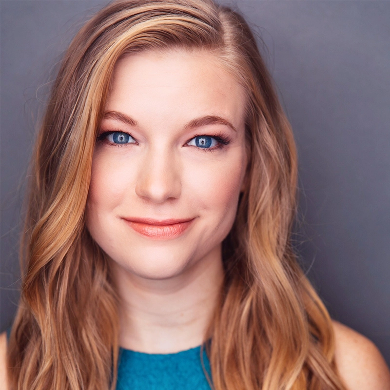 A young woman with long, wavy blonde hair and blue eyes smiles gently. She wears natural makeup with peach lipstick and a teal sleeveless top. The background is a plain, soft gray, and the lighting highlights her face.
