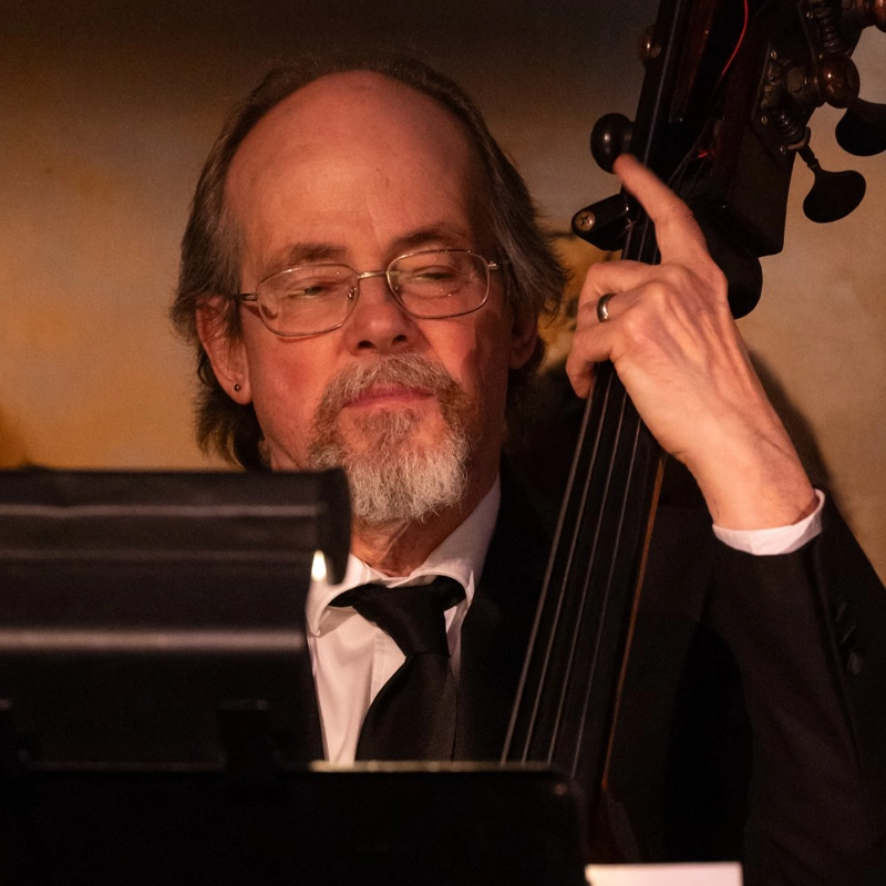 A middle-aged man with glasses, a short gray beard, and balding hair plays an upright bass. He is dressed in a black suit and tie. His expression is focused as he looks down at sheet music in front of him while performing.
