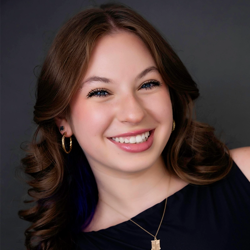 Arielle Ziff, a young woman with long, brown, wavy hair, smiles at the camera. She wears gold hoop earrings, a gold necklace, a sleeveless black top, and subtle makeup. The dark gray background makes her bright face stand out.