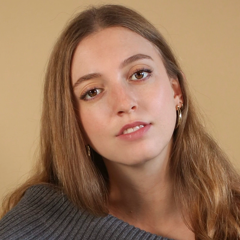 A young woman with long, light brown hair and hoop earrings looks directly at the camera. She is wearing a gray, off-the-shoulder knit sweater. The background is a plain beige color, and her expression is calm and relaxed.