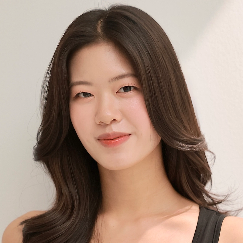 A young woman with long, wavy dark hair smiles gently at the camera. She is wearing a black sleeveless top and stands against a plain, light-colored background, with soft lighting highlighting her natural features.