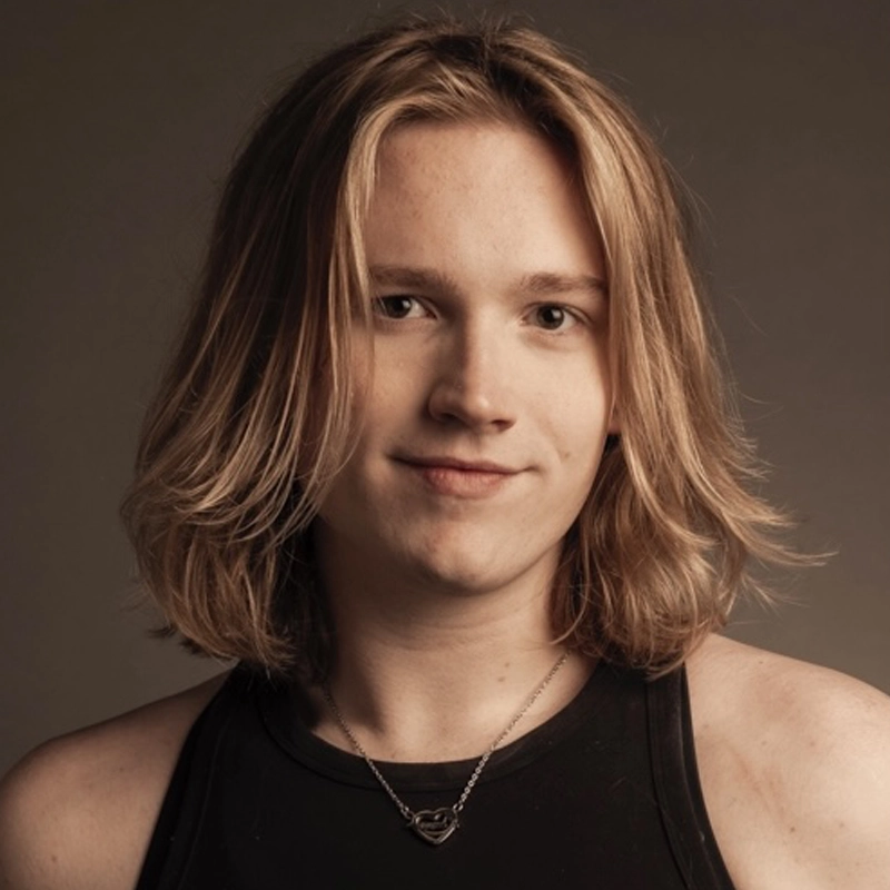A young person with shoulder-length blonde hair smiles gently at the camera. Hailee Ashton wears a sleeveless black top and a silver necklace with a small heart-shaped pendant. The softly lit, plain background creates a warm, neutral atmosphere.