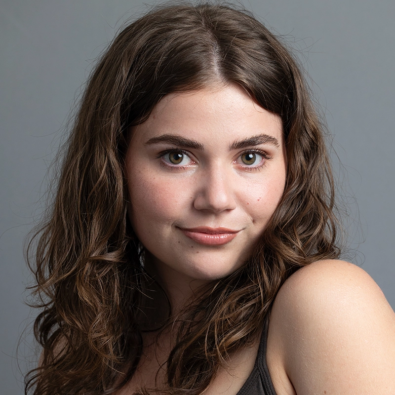 Arielle Ziff, a young woman with wavy brown hair and fair skin, smiles softly at the camera. She has hazel eyes, natural makeup, and wears a black sleeveless top against a plain gray background that highlights her expression.