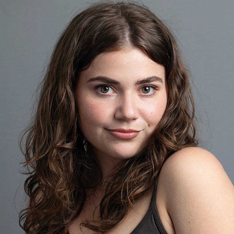 A young woman with long, wavy brown hair and fair skin poses against a plain gray background. She wears a dark tank top, looking directly at the camera with a gentle smile and bright green eyes. Her expression is confident and friendly.