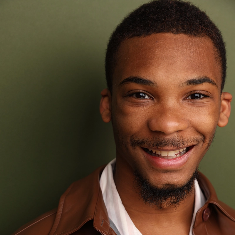 A young man with short hair and a goatee smiles warmly. He wears a brown jacket over a white shirt and stands against an olive green background. Only the right half of his face is visible, capturing a joyful, relaxed expression.