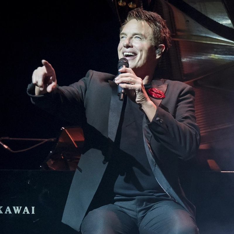 A man in a black suit with a red pocket square—John Barrowman—sits onstage holding a microphone, smiling and pointing outward. He appears to be performing, with a Kawai piano partially visible behind him under stage lighting.