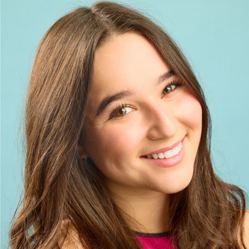 A young woman with long brown hair and glowing skin smiles at the camera. She has brown eyes, natural makeup, and is wearing a sleeveless pink top. The background is a solid light blue, creating a bright, cheerful atmosphere.