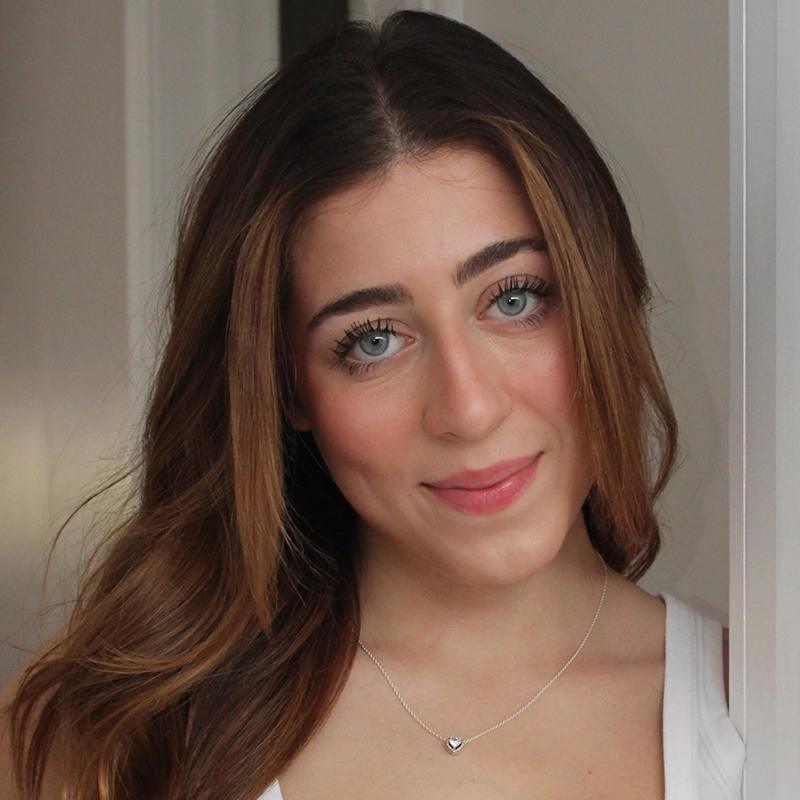 Bella Aboulhosn, a young woman with long, wavy brown hair, smiles gently. She has blue eyes, natural makeup, and wears a white top with a silver heart pendant necklace. She stands indoors against a neutral background with soft lighting.