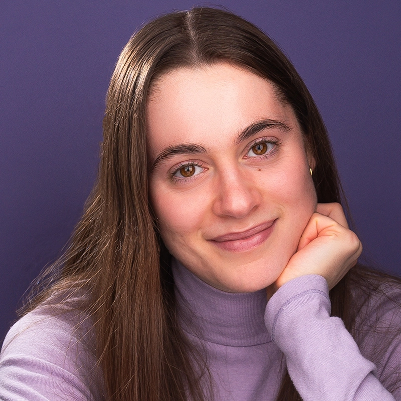 A young woman with long brown hair and fair skin smiles gently, resting her chin on her hand. She wears a light purple turtleneck and sits in front of a solid purple background. The lighting is soft, highlighting her relaxed and friendly expression.