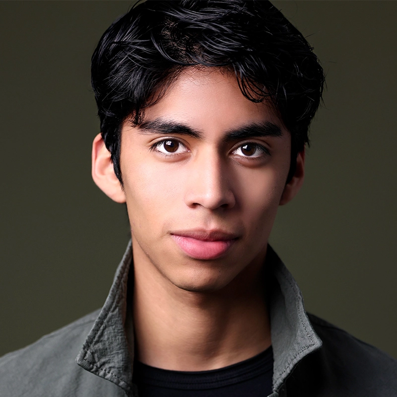 A young man with medium-dark skin and short, wavy black hair looks directly at the camera. He wears a dark jacket over a black shirt, his intense gaze reminiscent of portraits by Mary Grace Smith, set against a plain dark olive-green background.