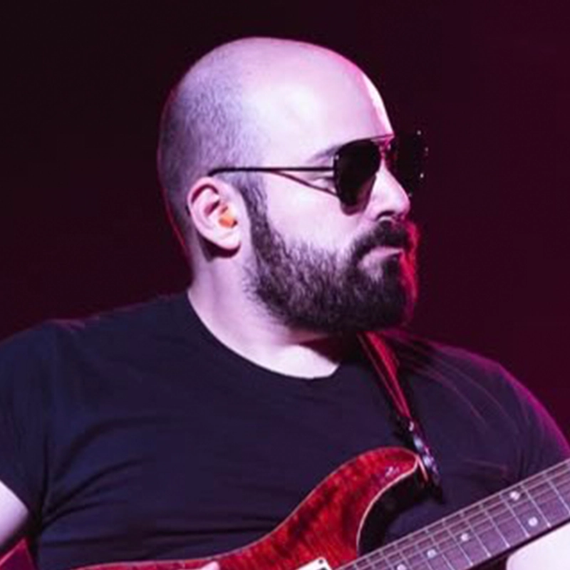 A bearded man with a bald head, wearing dark sunglasses and a black t-shirt, plays a red electric guitar on stage. Purple light bathes the scene as Ben Shanblatt delivers a moody concert atmosphere.
