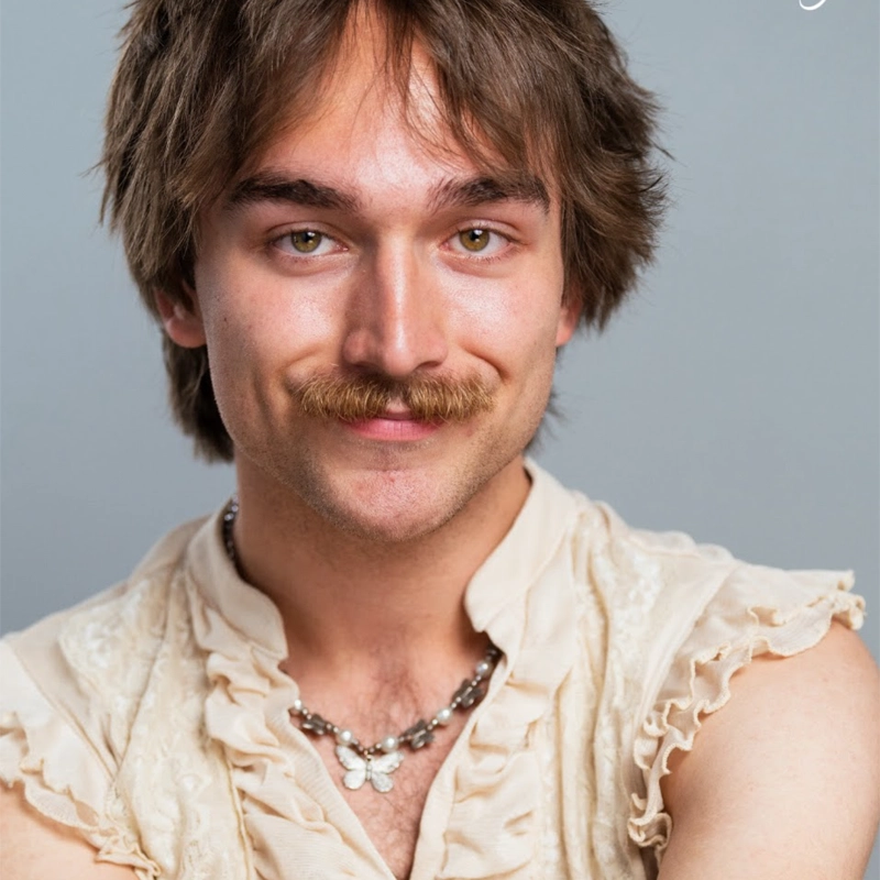 A young person with light skin, green eyes, and a thick mustache smiles gently at the camera. Michael Blanco wears a cream ruffled sleeveless blouse and a silver necklace with a butterfly pendant, set against a plain light gray background.