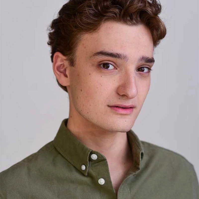 A young man with short, wavy brown hair wears an olive green button-up shirt. Jesse Brasler is facing slightly to the right, looking at the camera with a neutral expression. The background is plain and light-colored.