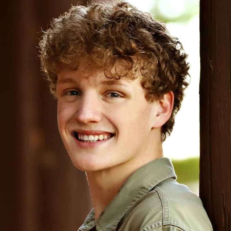 Devon Breese, a young person with curly light brown hair, smiles warmly at the camera. They are wearing a green collared shirt and leaning against a wooden post, with softly blurred greenery in the background.