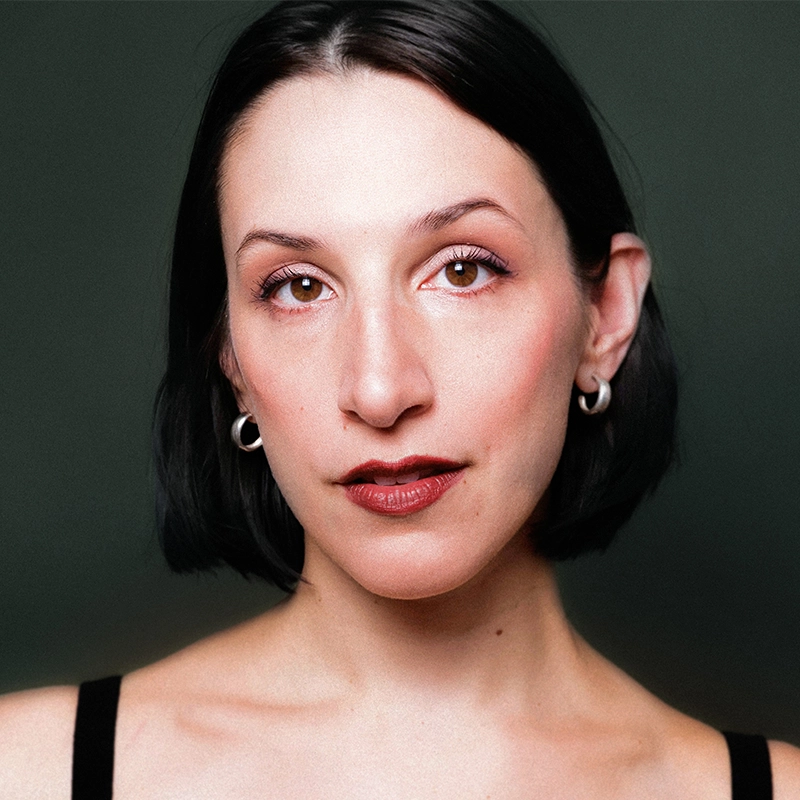 A woman with short, straight black hair and fair skin looks directly at the camera. She wears silver hoop earrings, dark red lipstick, and minimal makeup. The background is dark green, and she has a calm, confident expression.