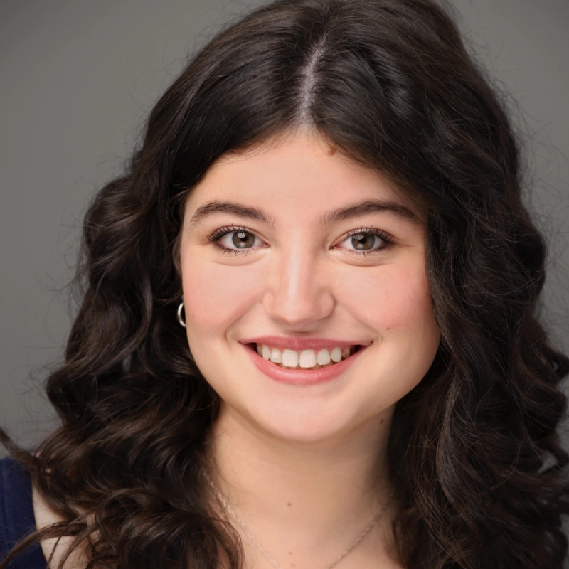 Stella Burnside, a young woman with long, dark curly hair, smiles at the camera. She has light skin, brown eyes, and wears natural makeup with subtle pink lipstick. She sports a dark top and a silver necklace against a plain gray background.