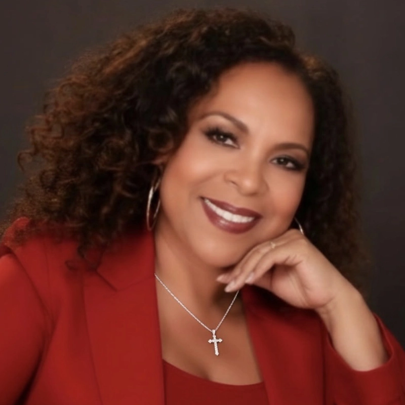 Deborah Burrell-Cleveland, with curly dark hair, smiles in a red blazer and top. She rests her chin on her hand, showing a silver cross necklace and hoop earrings. The dark, plain background highlights her warm expression.