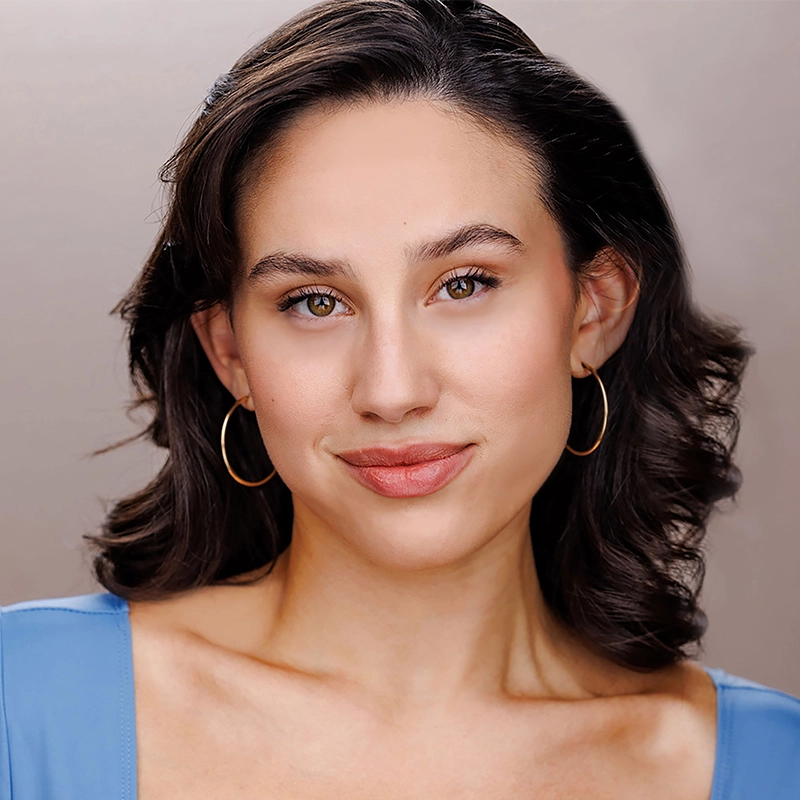 A young woman with wavy dark brown hair and gold hoop earrings smiles softly. She wears a light blue top with a scoop neckline. The background is a soft beige, keeping the focus on her natural, warm expression.