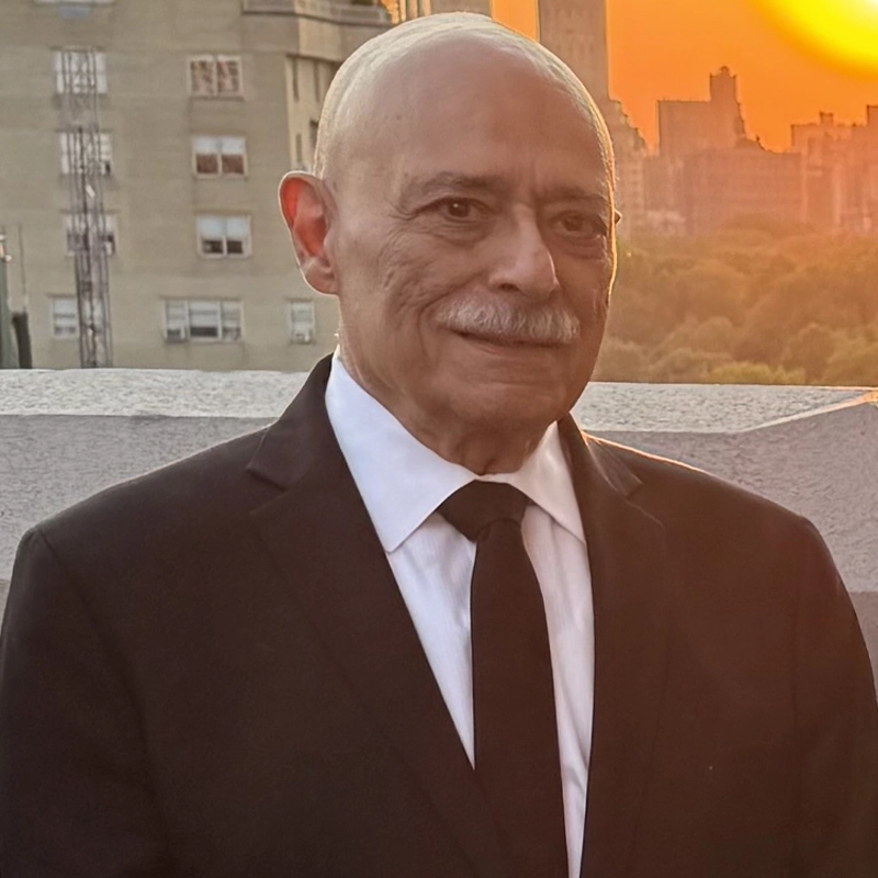 Ed Caccavale, an older man with a bald head and gray mustache, dressed in a black suit, stands on a rooftop at sunset. City buildings and trees fill the background as the orange sun sets low in the sky.