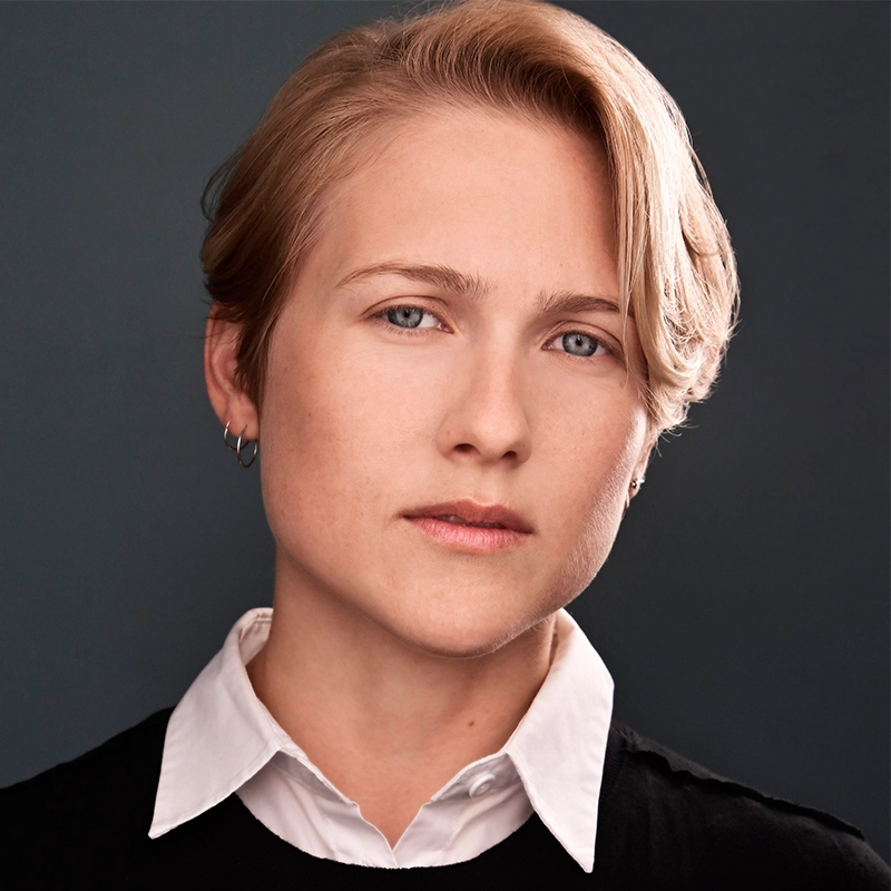 A person with short blonde hair and light skin looks into the camera with a neutral expression. They are wearing a white collared shirt under a black sweater and small hoop earrings. The background is plain and dark gray.