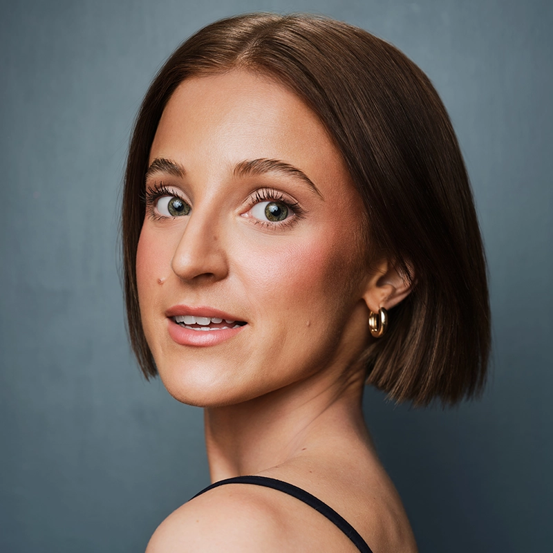 A young woman with fair skin and straight brown hair cut in a blunt bob poses in front of a gray background. Styled by Isaac Woofter, she has green eyes, rosy cheeks, gold hoops, and a black sleeveless top, looking over her shoulder with a soft smile.