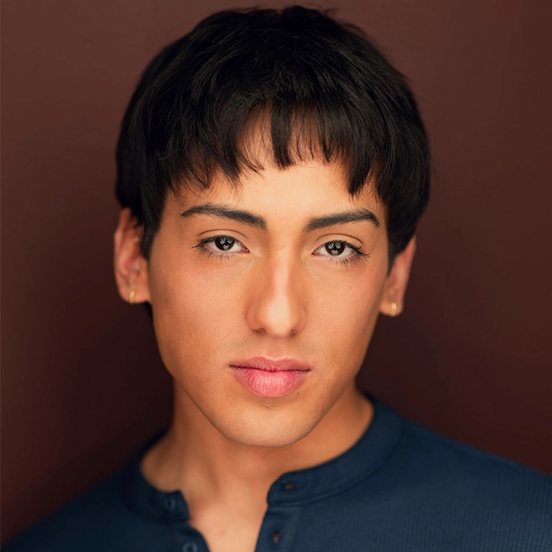 Arielle Ziff, a young person with short, dark hair and a straight fringe, stares directly at the camera. They have smooth skin, subtle makeup, light pink lips, small gold earrings, and wear a blue shirt against a solid dark brown background.