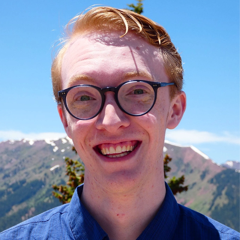 A smiling Shenandoah University BFA Musical Theatre student with short, reddish-blonde hair and glasses looks directly at the camera. They wear a blue collared shirt against a backdrop of a clear blue sky and sunlit, snow-capped mountains.