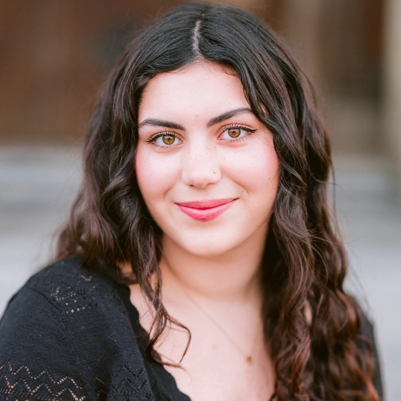 A young woman with long, wavy dark hair and hazel eyes smiles softly at the camera. Samara Castro wears a black top with subtle patterns and a delicate necklace. The background is softly blurred, drawing focus to her face.
