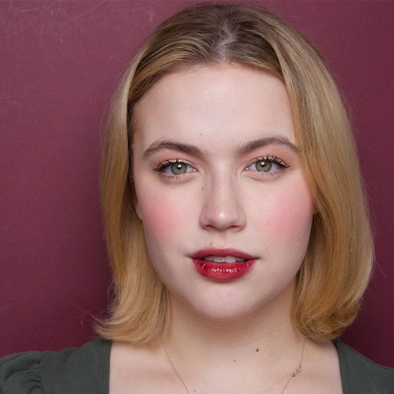 A woman with light skin and shoulder-length blonde hair stands against a maroon background. She has green eyes, subtle eye makeup, rosy cheeks, and glossy red lips. She is wearing a green top and a delicate gold necklace, gazing directly at the camera.