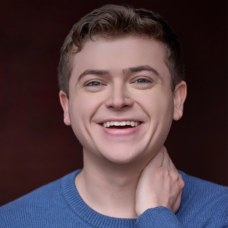 A young person with short, curly light brown hair smiles warmly, showing their teeth. They wear a blue sweater and gently touch their neck with one hand. The background is a dark, blurred brown, drawing focus to their expressive, happy face.