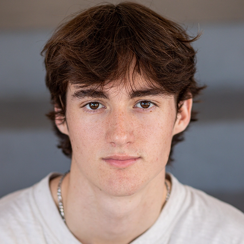 Connor DeRoche, a young man with medium-length brown hair and brown eyes, looks directly at the camera. He has a neutral expression, fair skin with freckles, and is wearing a white shirt and a silver chain necklace. The background is blurred with grayish tones.
